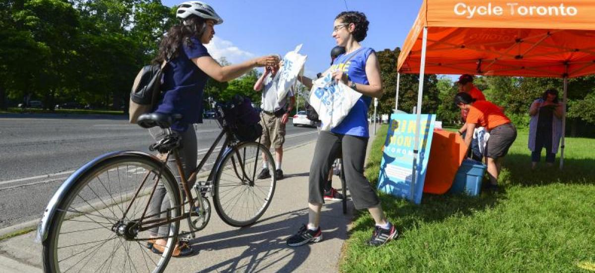 Commuter Station A smiling person hands a reusable bag filled with goodies to a person on a bike.