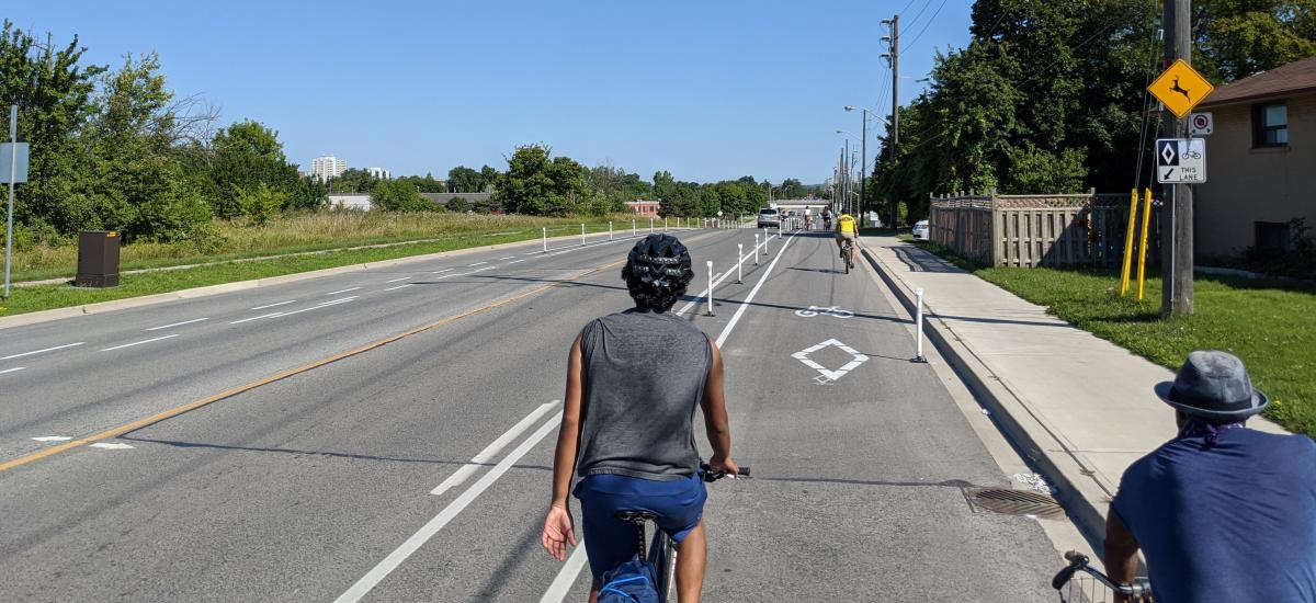 A wide road. No cars are in the two centre lanes. The two outer lanes are bike lanes and they have several people on bikes in them.