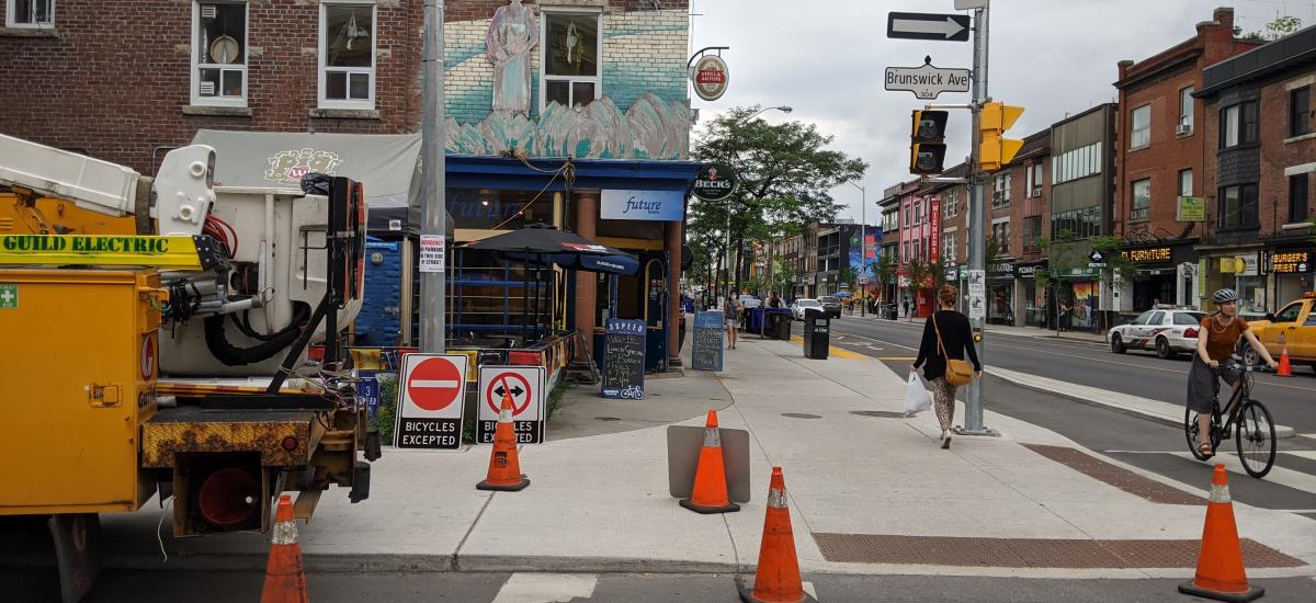 Photo of Bloor St at Brunswick Ave. In the foreground, an Electrical vehicle is parked and there are pylons surrounding it on the road and sidewalk. There are new signs on the sidewalk to be installed on the street for the planned contra-flow bike lanes o