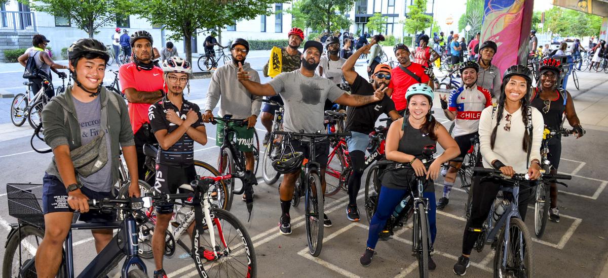 A diverse group of people pose with their bike. A man in the centre has his arms spread wide.