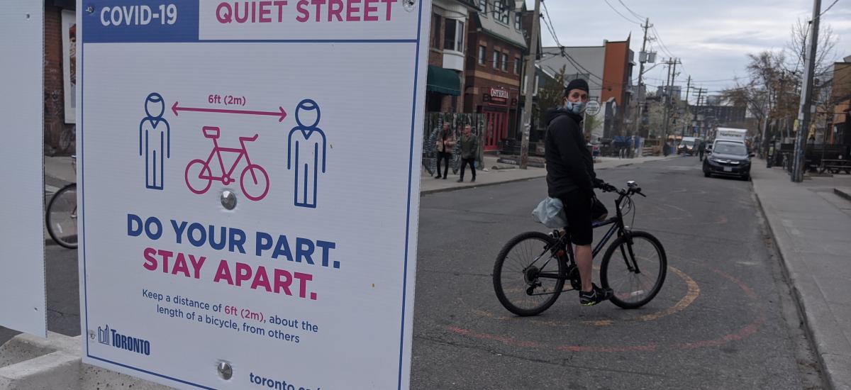 Person with a mask on a bike beside a sign that reads "Covid-19 Quite Street. Do your part. Stay Apart.