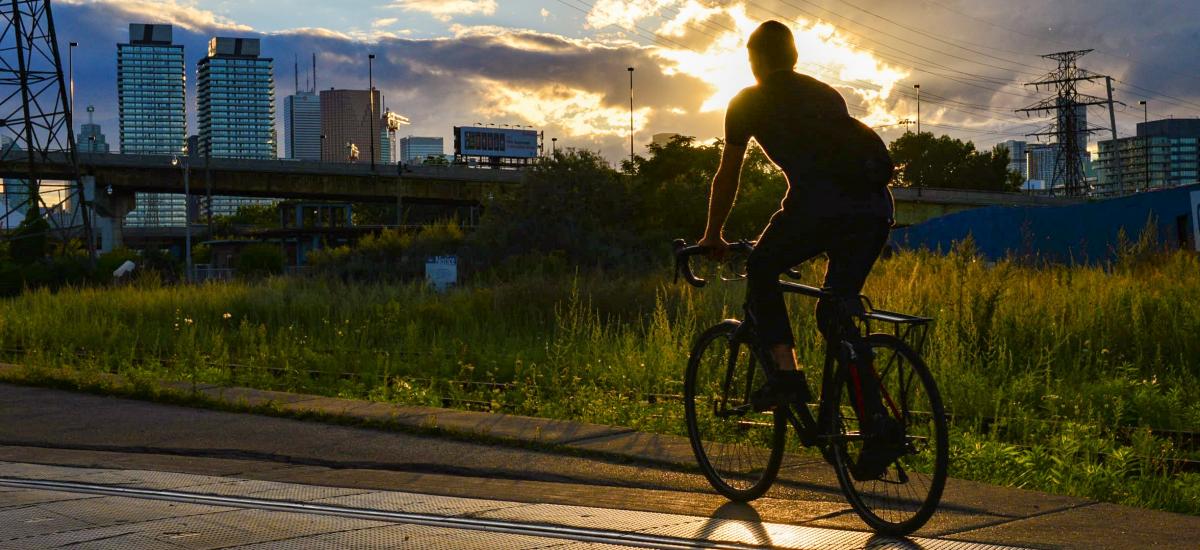 A person rides a bike over streetcar tracks in an industrial area. The setting sun outlines them leaving their shape a shadow in the foreground.