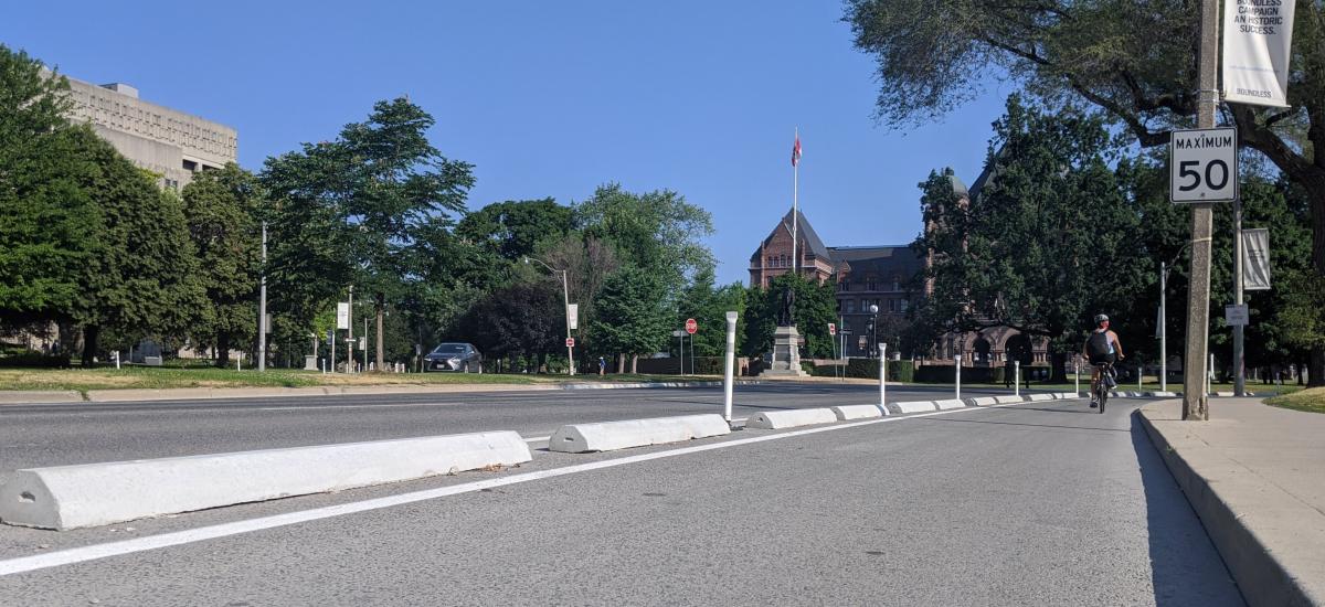 Concrete curbs and bollards separate a bike lane from a wide boulevard. A magnificent building is in the background