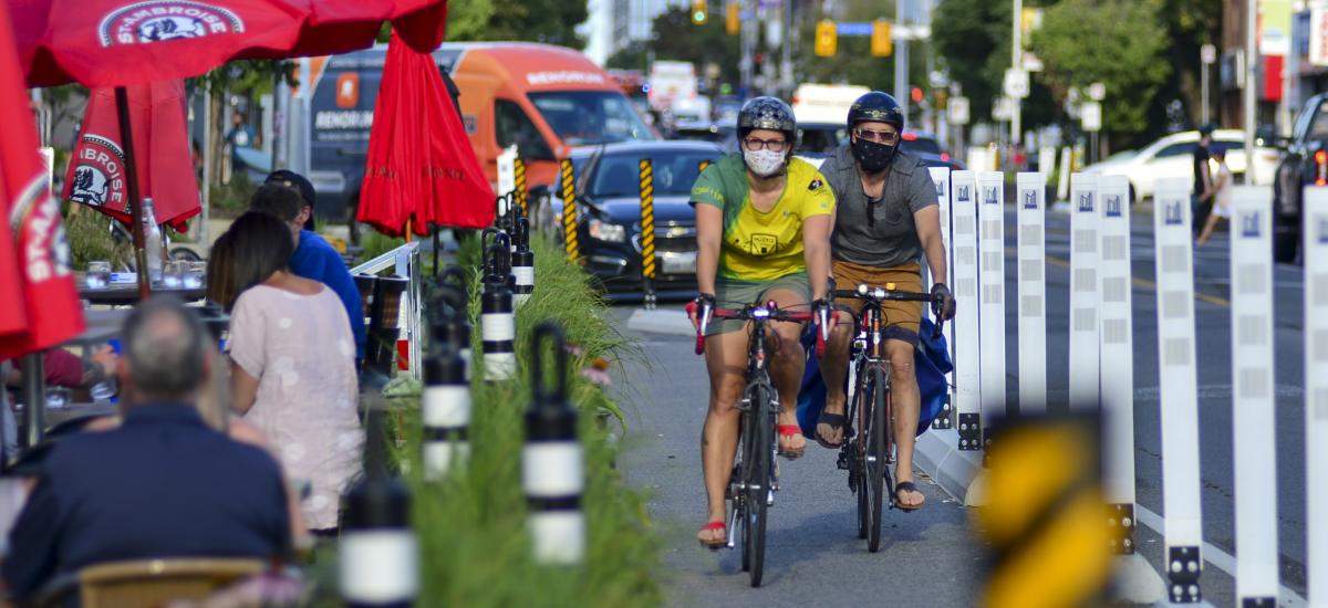 People wear masks as they ride bikes in a protected bike lane beside street-level patios