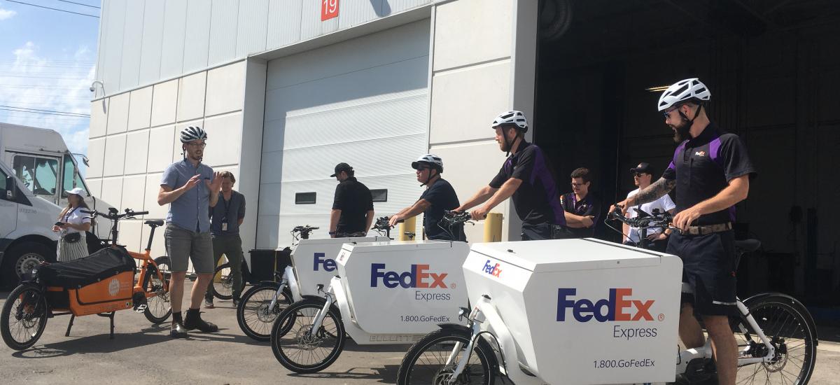 Several FedEx cargo bikes with riders are lined up next to a Cycle Toronto cargo bike. A person in plain clothes gives instructions