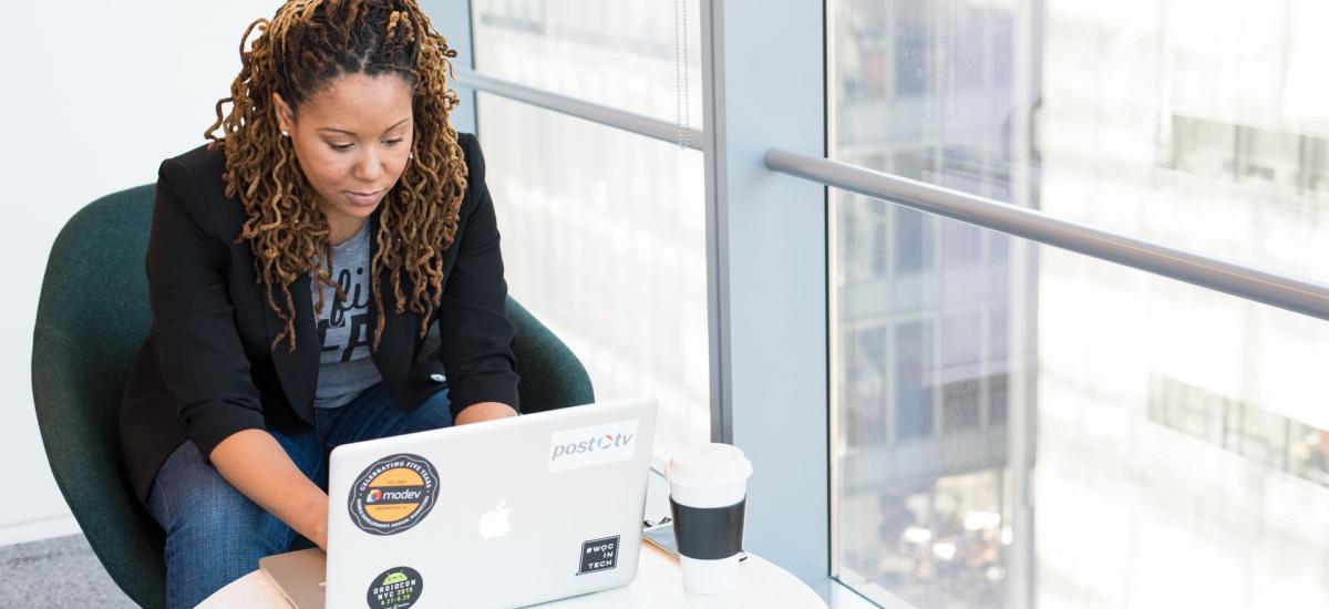 A woman works on a laptop. She is sitting at a table in a bright area next to a row of widows.