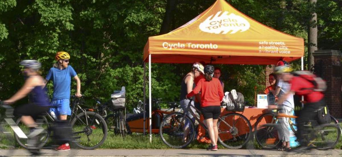 An orange tent with "Cycle Toronto" written on it is set up in a park. People with bikes ride by and huddle around it.