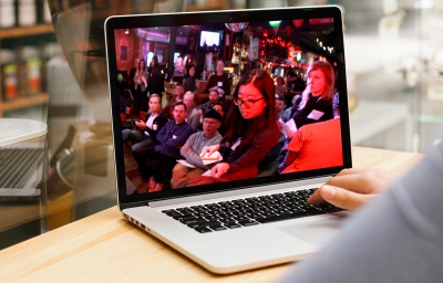 A laptop shows a photo of a Cycle Toronto member casting their vote at the 2019 Cycle Toronto Annual General Meeting. In the background, a group of people look on.