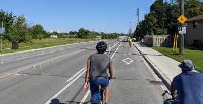 A wide road. No cars are in the two centre lanes. The two outer lanes are bike lanes and they have several people on bikes in them.