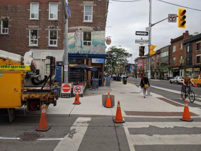 Photo of Bloor St at Brunswick Ave. In the foreground, an Electrical vehicle is parked and there are pylons surrounding it on the road and sidewalk. There are new signs on the sidewalk to be installed on the street for the planned contra-flow bike lanes o