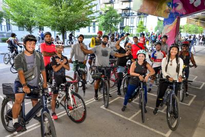 A diverse group of people pose with their bike. A man in the centre has his arms spread wide.