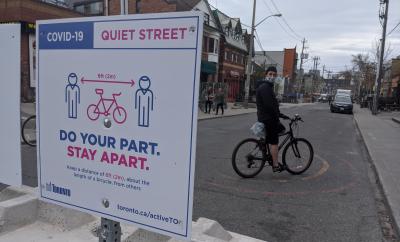 Person with a mask on a bike beside a sign that reads "Covid-19 Quite Street. Do your part. Stay Apart.