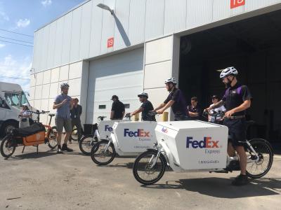 Several FedEx cargo bikes with riders are lined up next to a Cycle Toronto cargo bike. A person in plain clothes gives instructions