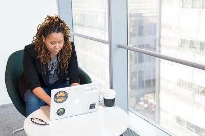 A woman works on a laptop. She is sitting at a table in a bright area next to a row of widows.