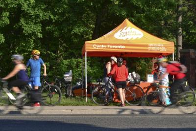 An orange tent with "Cycle Toronto" written on it is set up in a park. People with bikes ride by and huddle around it.