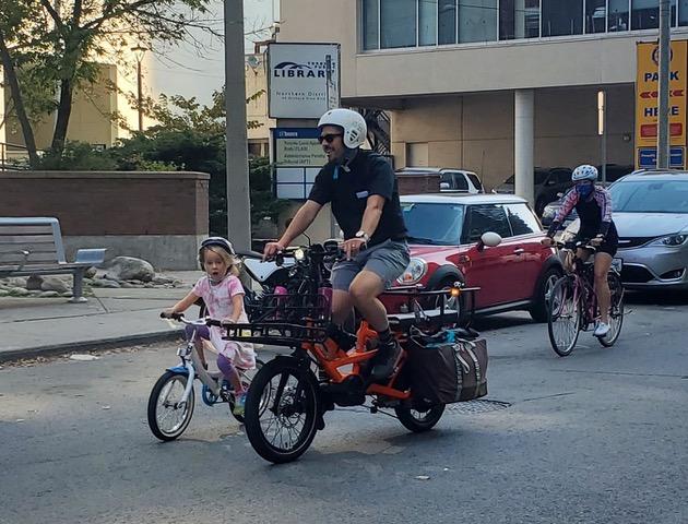 A man rides a packed cargo bike next to a very young person that is also riding a bike.