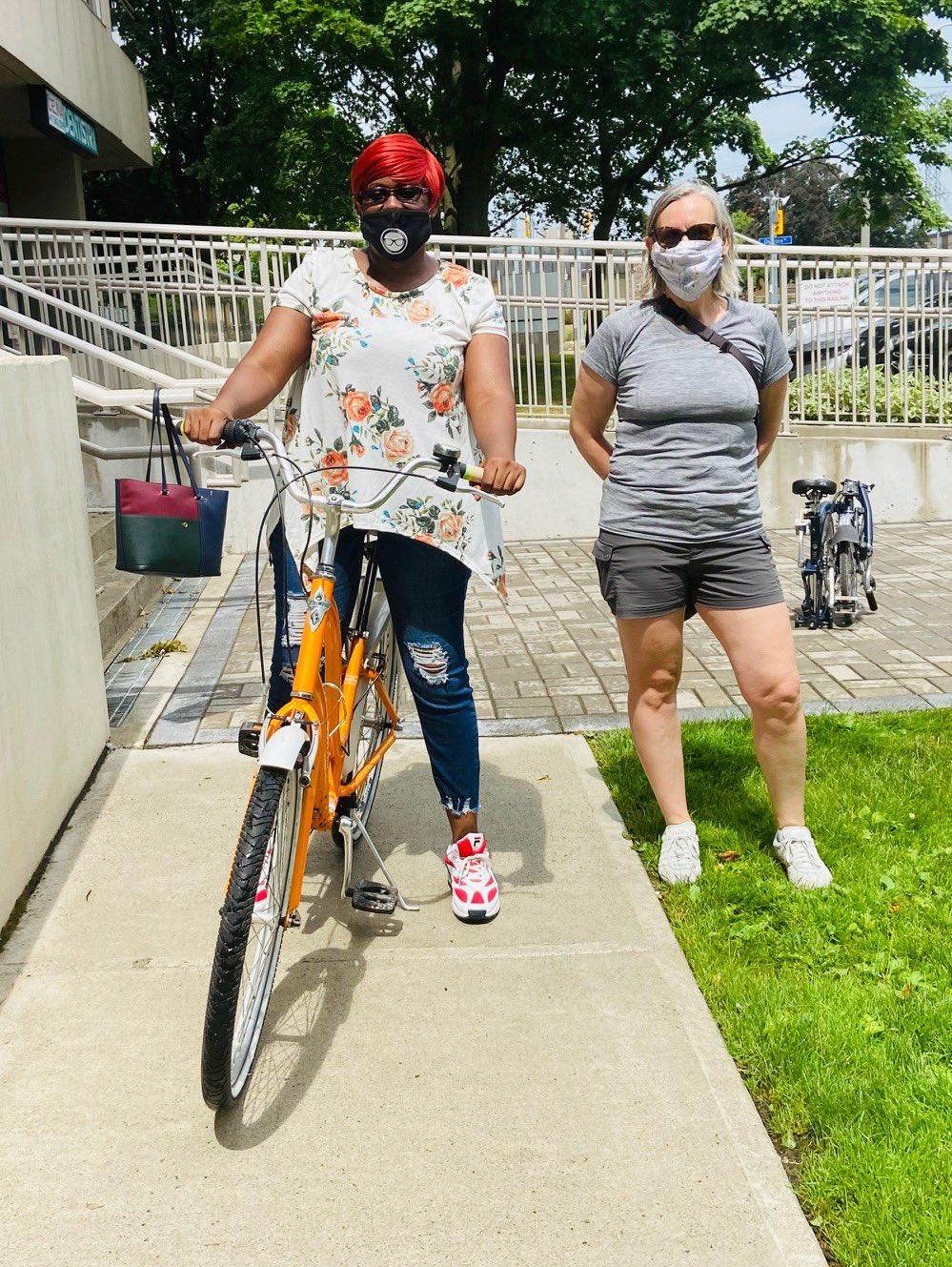 Two women with masks on pose. The woman on the left stands over a new looking orange bike.