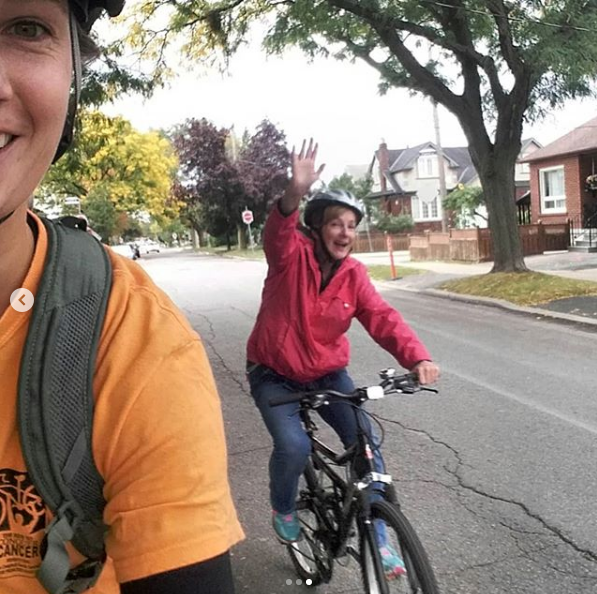 A person takes a selfie as another rides past while waving