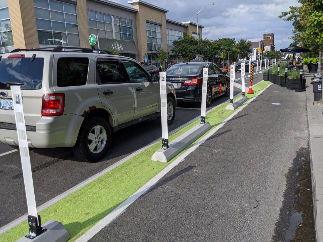 Destination Danforth project shows a bike lane on the right and parked cars on the left. Parked cars are separated from the bike lane with green paint, curbs, and bollards mounted on top of the curb.