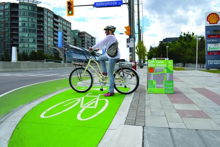 A person waits, sitting on their bicycle, to cycle through an intersection. They wait in a section of the road that has a bright green bike box painted, a location intended to be used to make a left-hand turn on a bicycle.