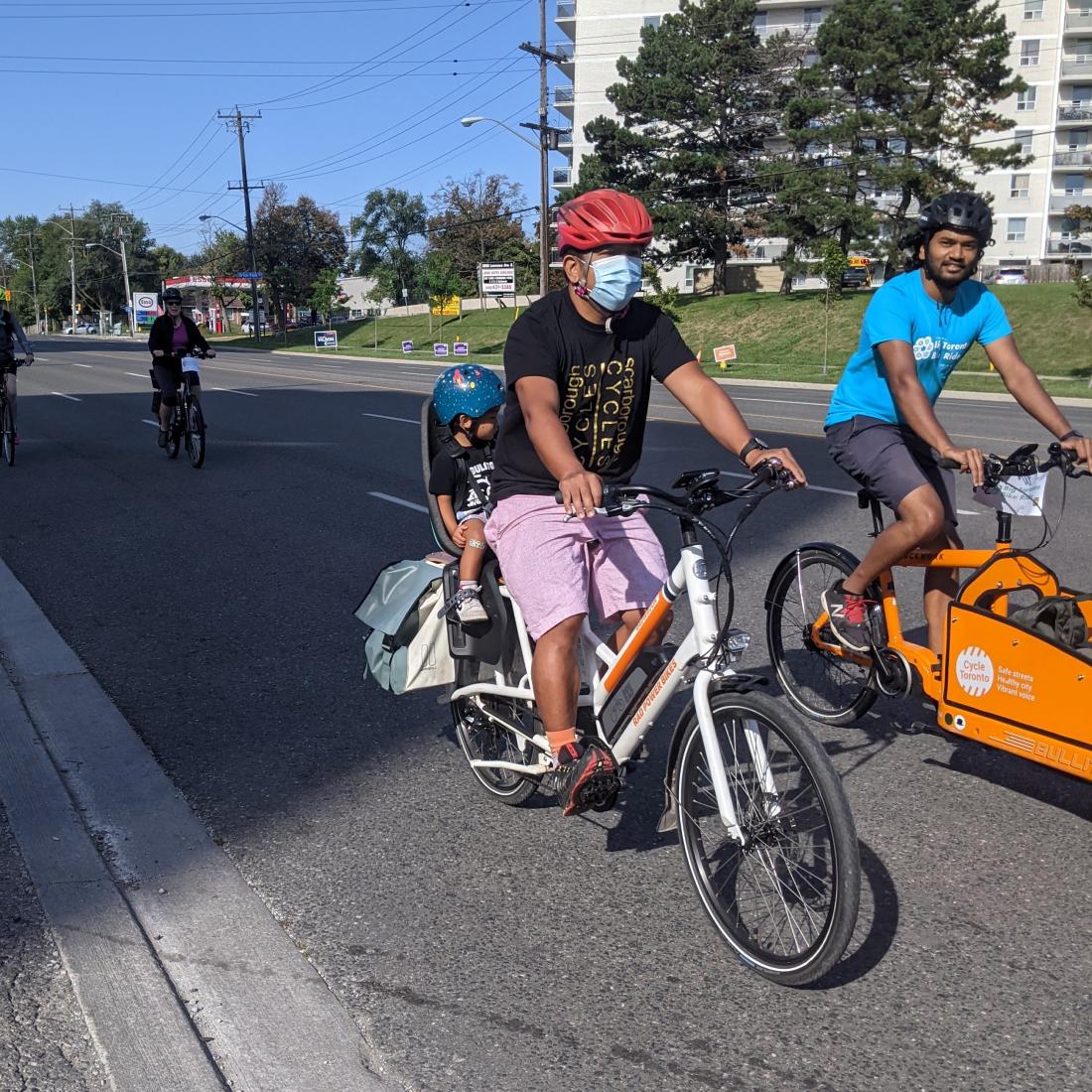 A man with a mask rides a bike with his child on the the back. 