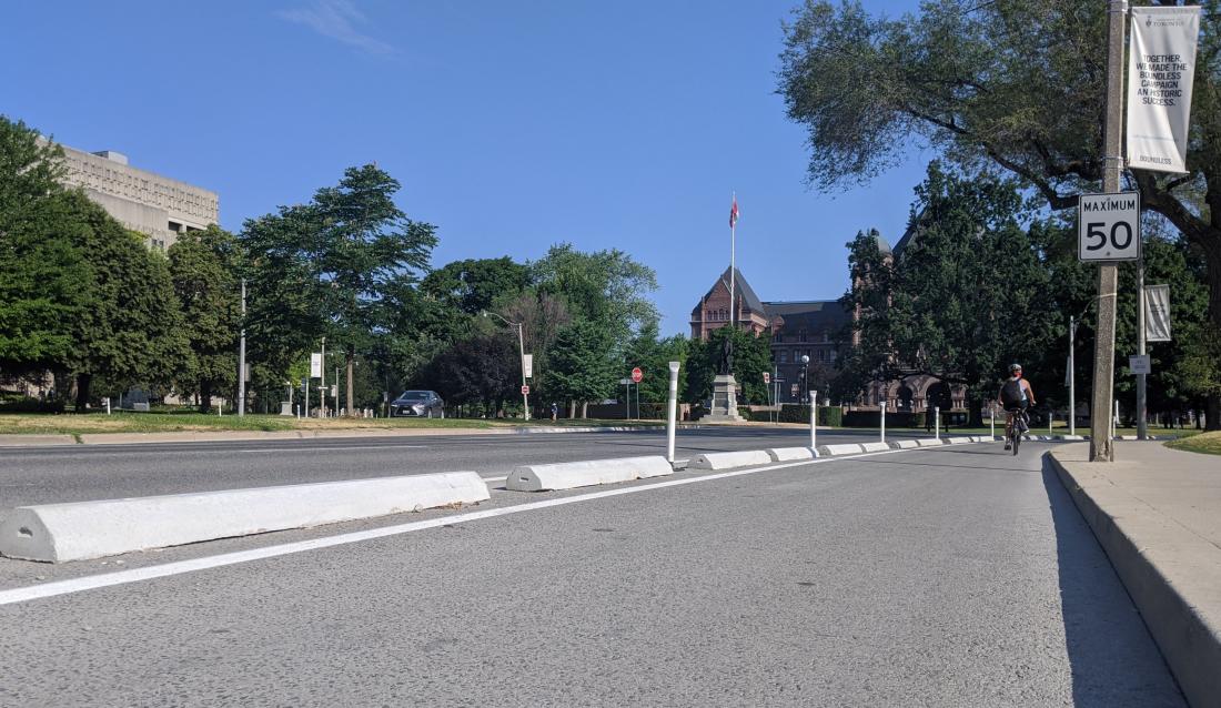 Concrete curbs and bollards separate a bike lane from a wide boulevard. A magnificent building is in the background