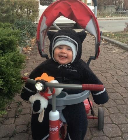 Violet Perry sits, smiling in a tricycle. She is bundled up for winter and has a black snowsuit and hat on.