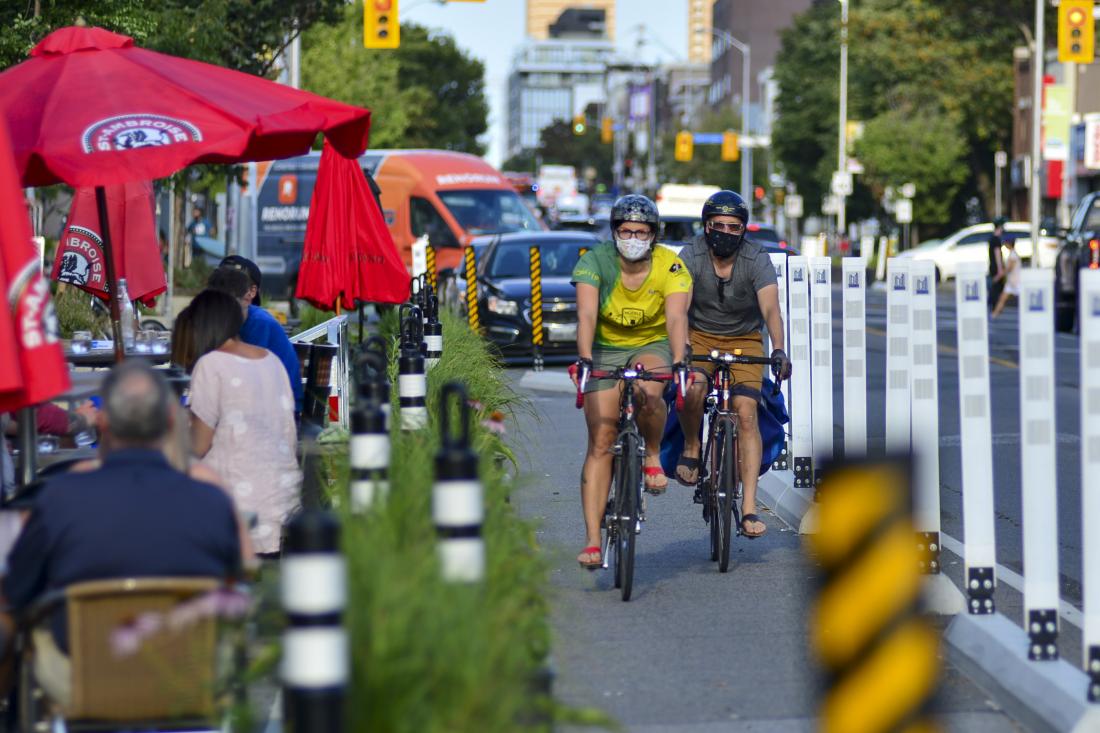 People wear masks as they ride bikes in a protected bike lane beside street-level patios