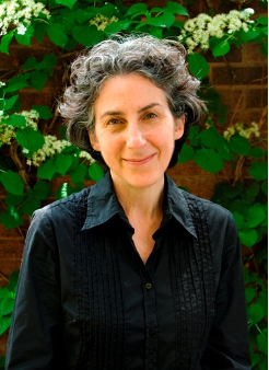 Julia Langer poses in front of a brick wall and greenery. Julia smiles